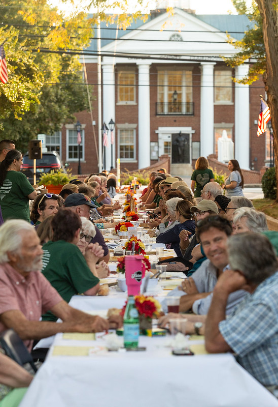 Image of 2024 Longest Table event. Many people sitting at a long white table talking and eating.